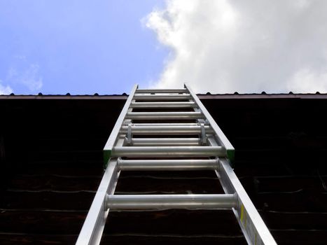 A silver ladder leans against a roof with cloudy sky.