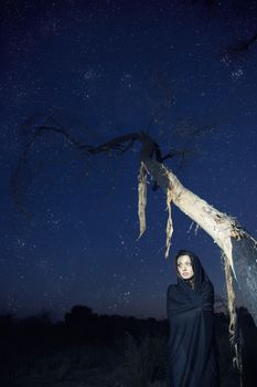 Female witch outdoors near the dead tree in deep night with stars in the sky