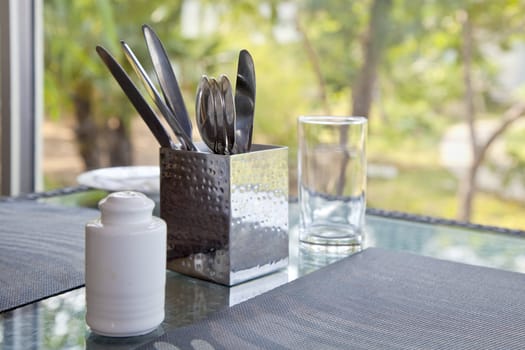 Horizontal capture of a dinner table waiting to be properly set at a picture window looking onto a garden. Place mats, cutlery knife and spoon, pepper pot and water glass