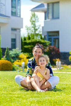 Happy girl with her mother sitting on the lawn in front new cottage. Vertical view