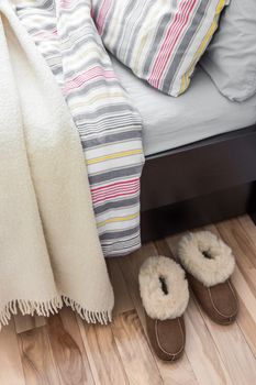 Cozy slippers on the floor, near a bed with striped bed linen.