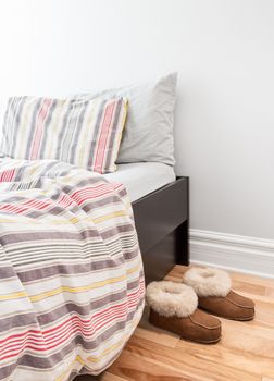 Warm cozy slippers near a bed with striped bed linen.