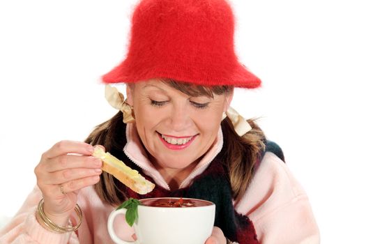 Middle aged woman dips a slice of bread in a mug of soup.
