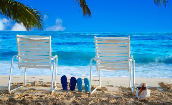 Two white Beach chairs under palm leaves by the ocean, with flip flops and seashell with sunglasses.