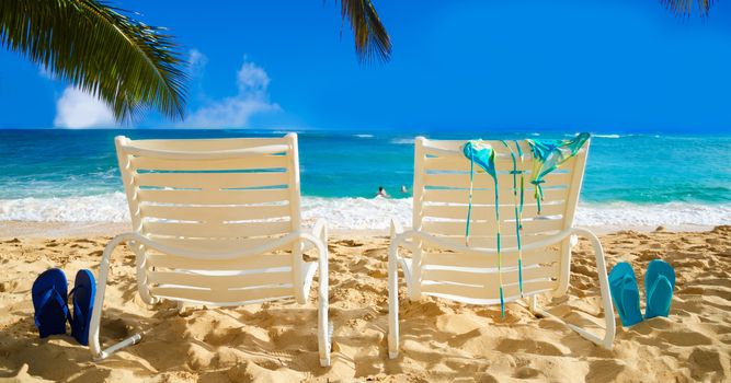 Two white beach chairs under palm leaves by the ocean, with bikini and flip flops and couple in the ocean on background