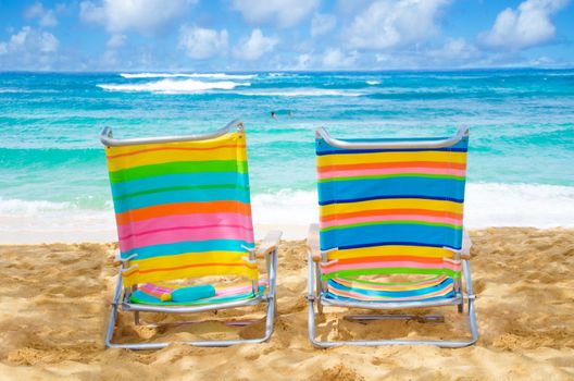 Two colorful beach chairs under by the ocean with sunscreen, with couple in the ocean on background