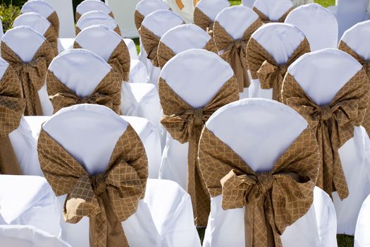 Chairs with white linen and decorative bows placed out on grass for a reception
