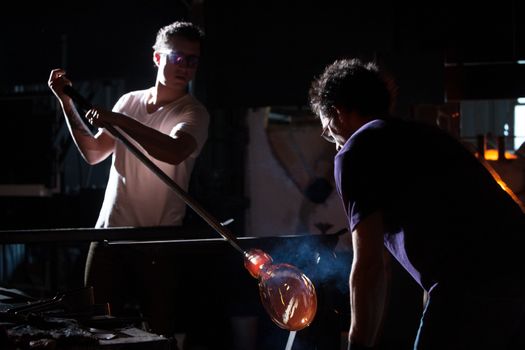 Two men working with hot glass vase in dark studio