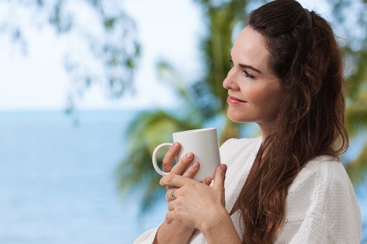 Close-up portrait of a beautiful woman enjoying her morning coffee or tea on a tropical balcony.