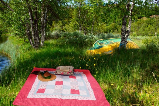 A pink patchwork blanket, strawhat, pillow and a book under the trees by the river
