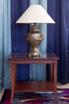 A living room lamp on a side table placed on a carpet and in front of a window with striped curtains