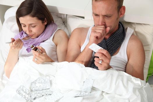 Husband and wife with the flu feeling very sorry for themselves as they sit up against the pillows in bed coughing and taking medication