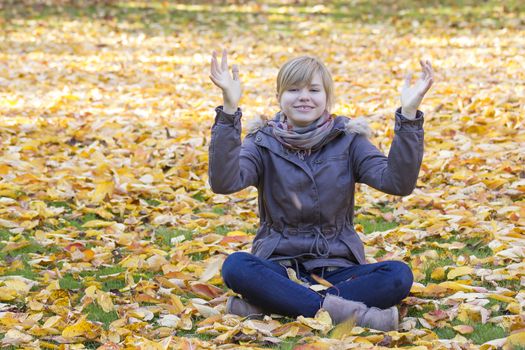 smiling girl sitting on fallen leaves in autumn park and rising up hands 