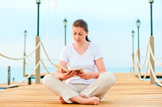 Young girl reading a book while sitting on a pier barefoot