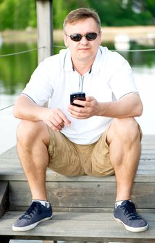 Mature man with sunglasses and smartphone sitting at pier