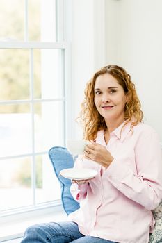 Smiling caucasian woman relaxing on couch by window holding cup of coffee
