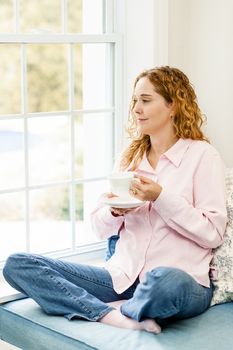 Smiling caucasian woman relaxing on couch by window holding cup of coffee