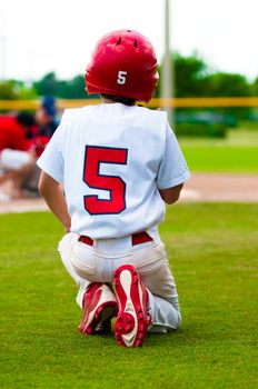 Baseball boy kneeling on the ground while an injured player is on the field.