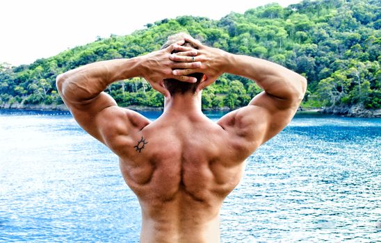 Muscular bodybuilder facing the sea with hands behind his head showing back, shoulders and arms