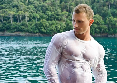 Attractive young bodybuilder by the sea with wet shirt on, serious expression