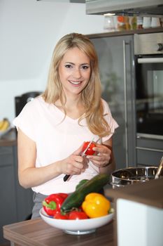 Smiling beautiful young housewife preparing a meal in the kitchen cutting a tomato to put into a saucepan on the stove