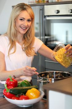 Smiling pretty young blond housewife preparing pasta in the kitchen tipping noodles into a saucepan of boiling water on the stove