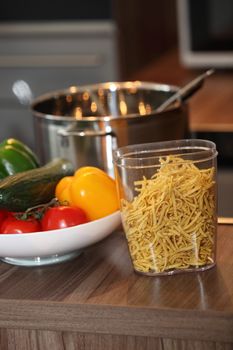 Fresh ingredients for pasta on a kitchen counter with a container of noodles and a bowl filled with fresh peppers and vegetables