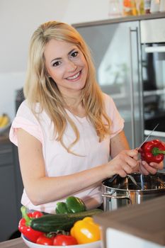 Smiling beautiful young woman preparing a meal standing in the kitchen slicing a fresh red bell pepper over a saucepan on the stove