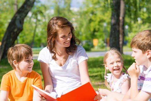 children and teacher reading book together in the summer park