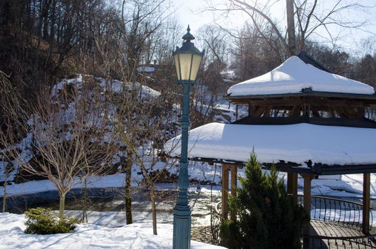 wooden bower roof covered with snow and retro lighting lamp pole on river water bank waterside in winter park.