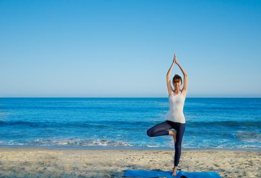 Young pretty woman practicing yoga on the beach by the ocean 