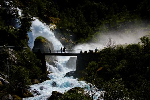 Tourits as dark silhouettes on the bridge at Briksdalsbreen watching the waterfall