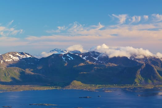 Scenic coastline of Lofoten islands in Norway with small fishing villages on the coast and snowy peaks covered by scattered clouds above them