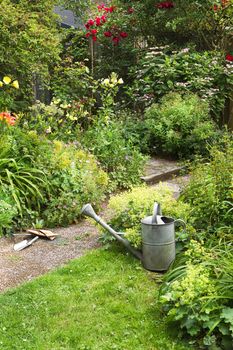 Watering-can and garden tools for cleaning blooming cottage-style garden on summer morning - vertical