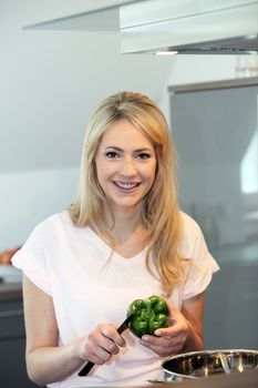 Beautiful woman slicing a green pepper with a knife while standing over a large metal pot on the stove in her kitchen