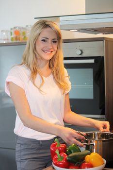 Pretty blond woman preparing a meal standing in the kitchen holding a pot over the stove alongside a colourful bowl of fresh vegetable ingredients