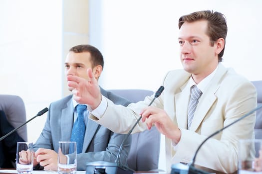 Image of two businesspeople sitting at table at conference speaking in microphone