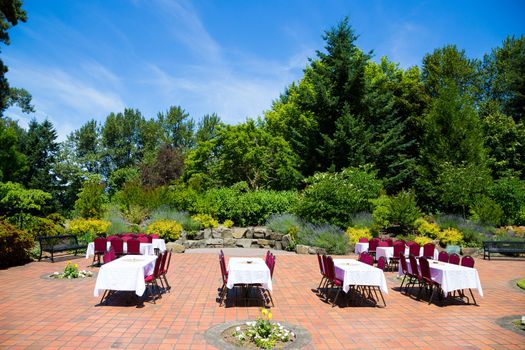 Some tables and chairs are set up ready for a wedding reception as soon the ceremony is finished outdoors in Oregon at a park. The sky is very blue and the patio is surrounded by nature and trees.