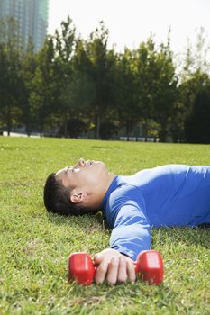 Young Athletic Man Lying Down in Park with Dumbbells, Vertical