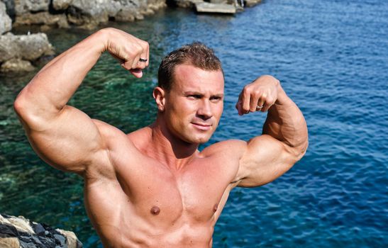 Double biceps pose for young, attractive bodybuilder by the sea showing muscular arms