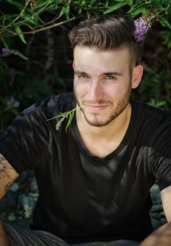 Attractive, smiling young man outdoors with flower between his lips, looking in camera