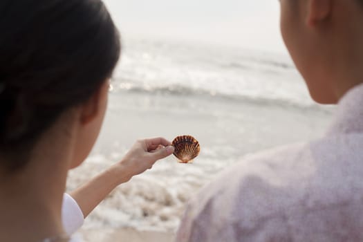 Young couple standing and looking at seashell on the beach, over the shoulder view