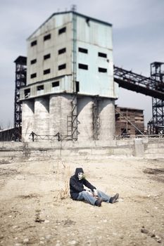 Alone man in gas mask sitting at the single plant. Industrial factory at the background. Shallow depth of field due to the tilt/shift lens for movie effect