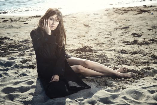 Sad woman in the black coat sitting on the sand at the autumn beach. Natural light and colors