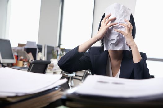 Young businesswoman sitting at desk covering her face with a paper