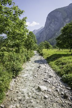 Picture of river in Austrian Alps with grey stones, meadow, trees, mountains and blue sky