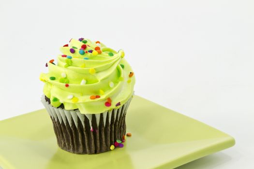 Colorful sprinkles on green frosting of chocolate cupcake on square plate with white background