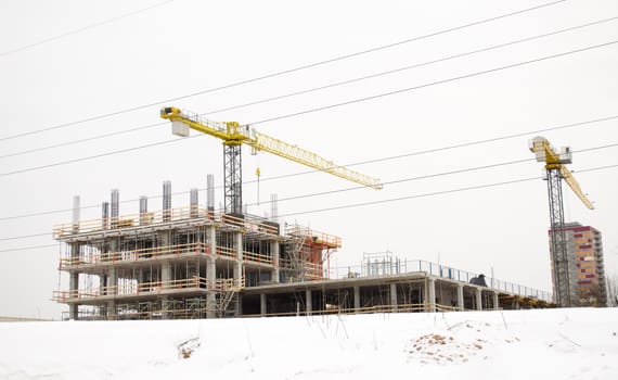 high building under construction and cranes on background of cloudy sky and winter snow.