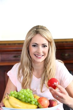 Smiling beautiful young blond woman with a bowl of healthy fresh fruit holding an apple in her hand