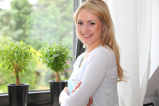 Beautiful young blond woman looking at the camera with a lovely smile standing in front of a view window overlooking a lush garden with topiary trees on the windowsill